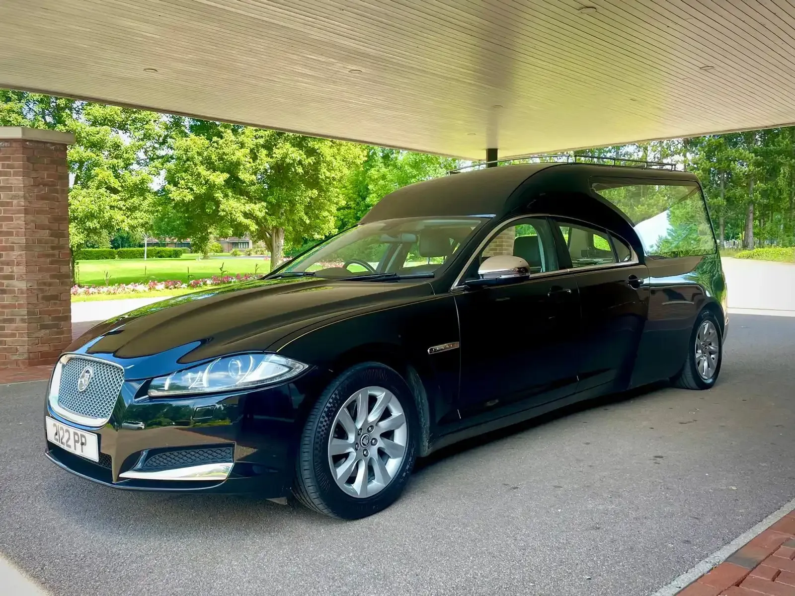 Black Jaguar hearse parked under a canopy, in front of greenery.