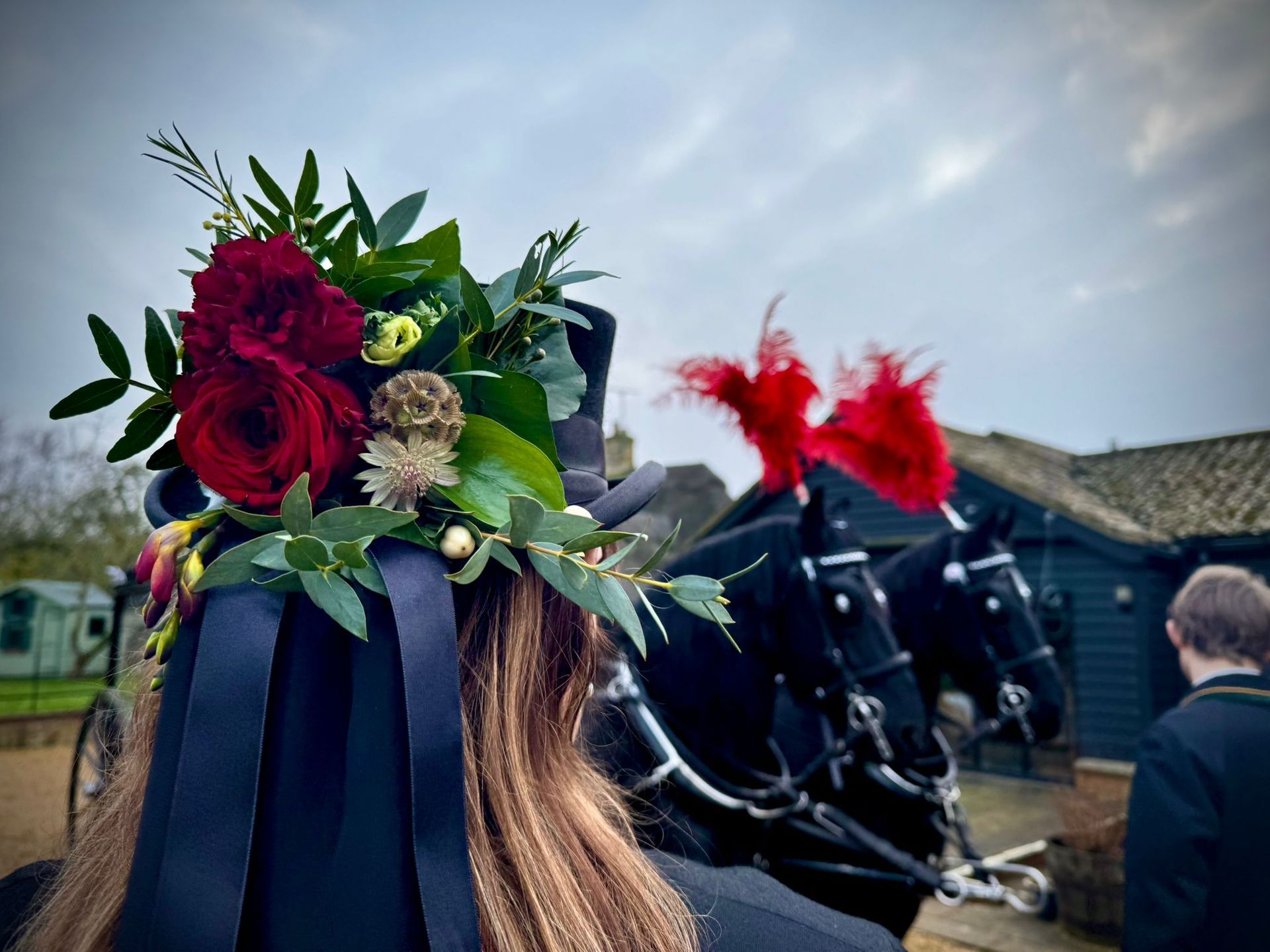 Person with floral headdress, dark ribbons, and horses with red plumes in front of a building.