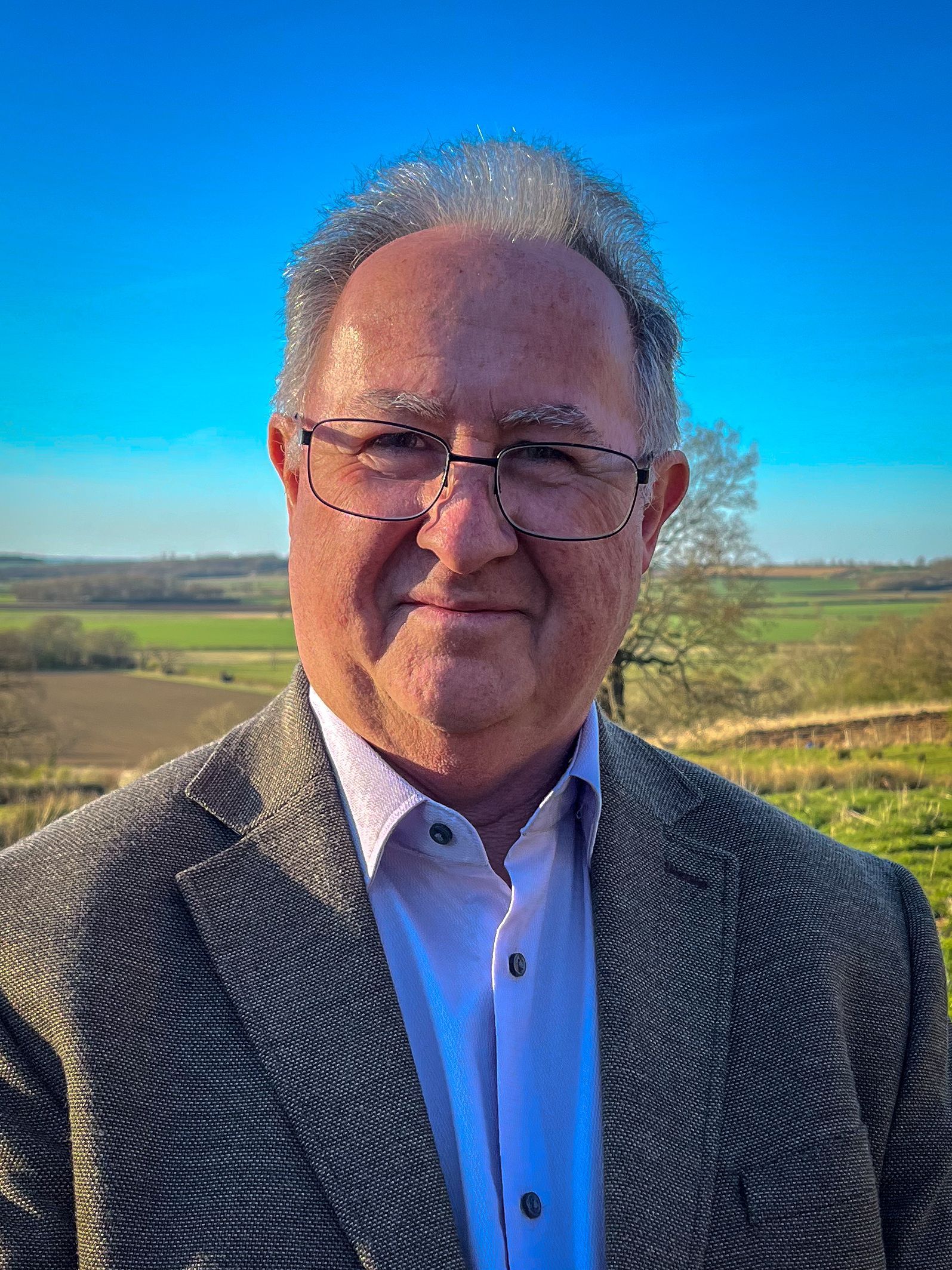 Man in glasses and blazer smiles outdoors, fields and sky in background.