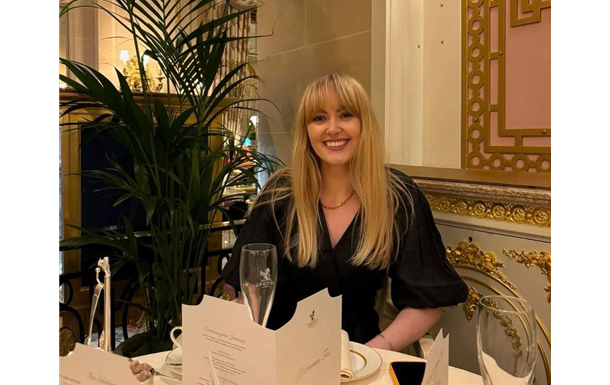 Smiling woman seated at a restaurant table with a wine glass, in a gold-trimmed dining room