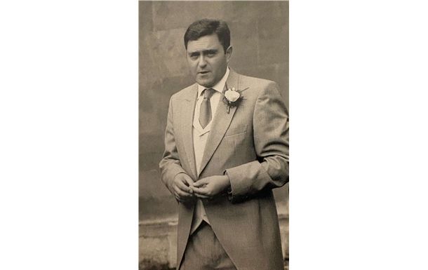 Sepia-toned portrait of a man in a suit standing with hands clasped, wearing a boutonniere.