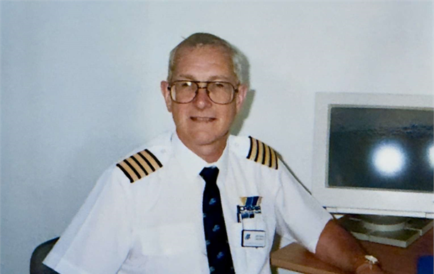 Man in white uniform shirt with epaulets sits beside a computer in an office.