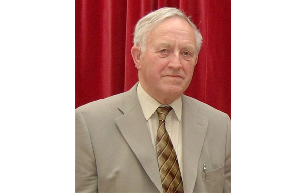 Portrait of a man in a beige suit and patterned tie against a red curtain background