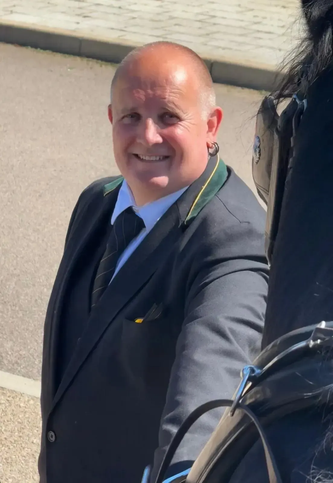 Smiling man in a dark suit and tie standing outdoors in sunlight, taking a selfie.