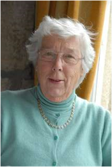 Elderly woman in a turquoise sweater and gold necklace, smiling by a window with curtains.