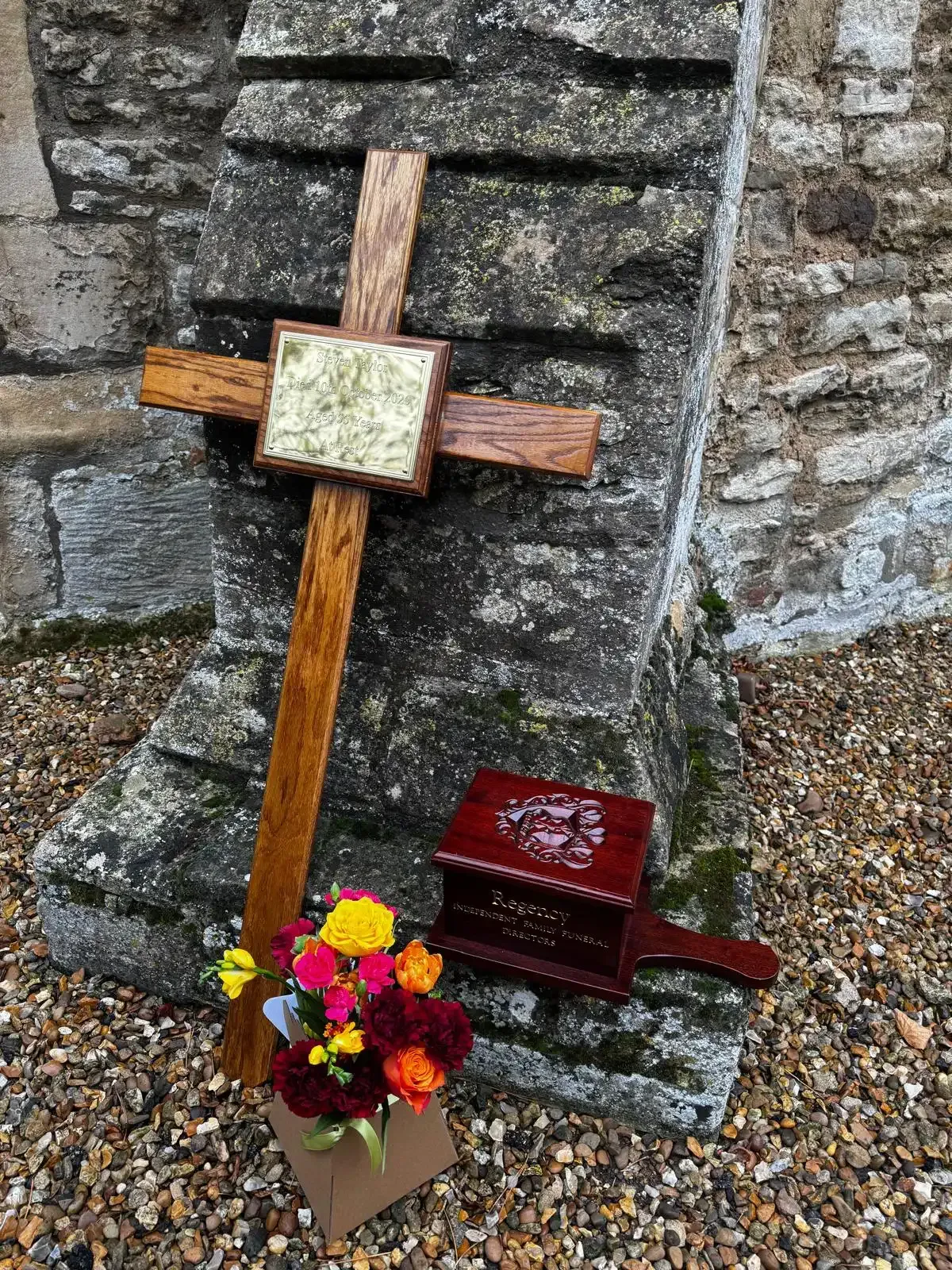 Wooden cross and memorial box with flowers against a stone wall.