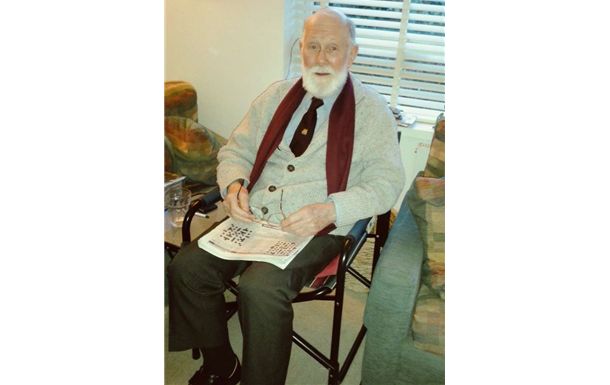 Man seated in a chair holding an open newspaper, wearing a light jacket and red vest, indoors near window blinds