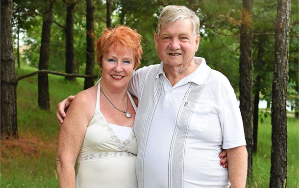 Couple smiling outdoors among trees, standing close together in light-colored clothing.