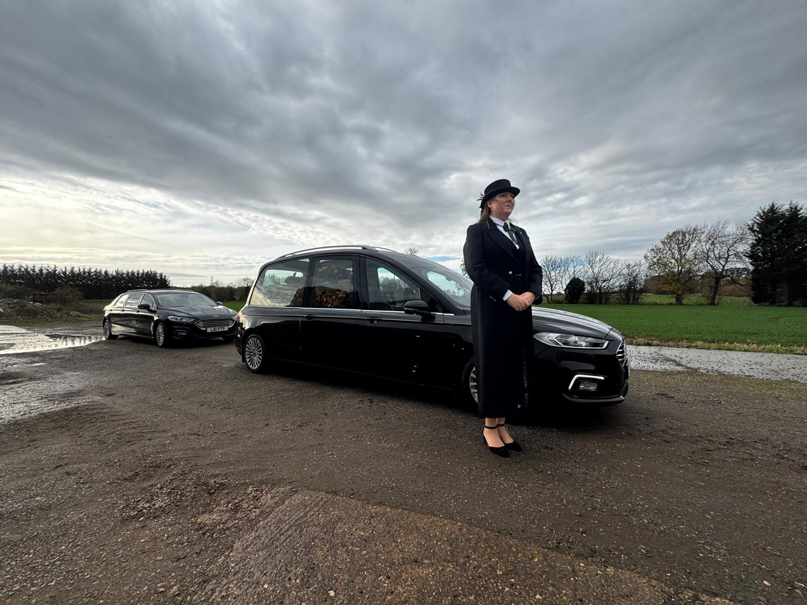 Woman in black suit stands by black car on a muddy road with another car in the background. Overcast sky.