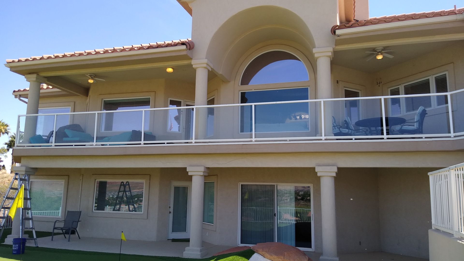 Two-story beige house with arched balcony, glass railing, and ground-floor patio under a clear sky with freshly cleaned glass