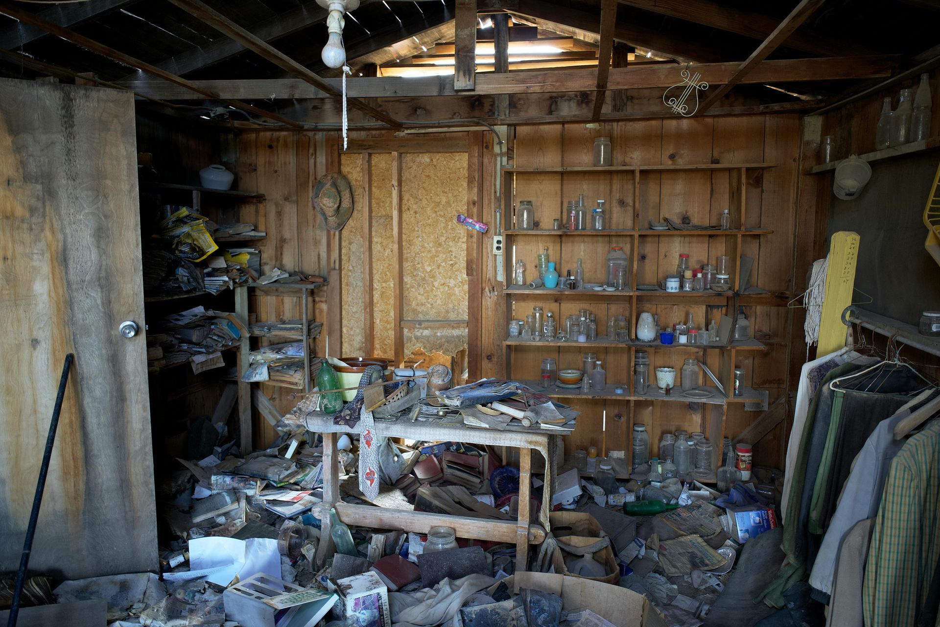 Cluttered, damaged room with broken furniture, scattered debris, shelves, and clothes hanging on the right