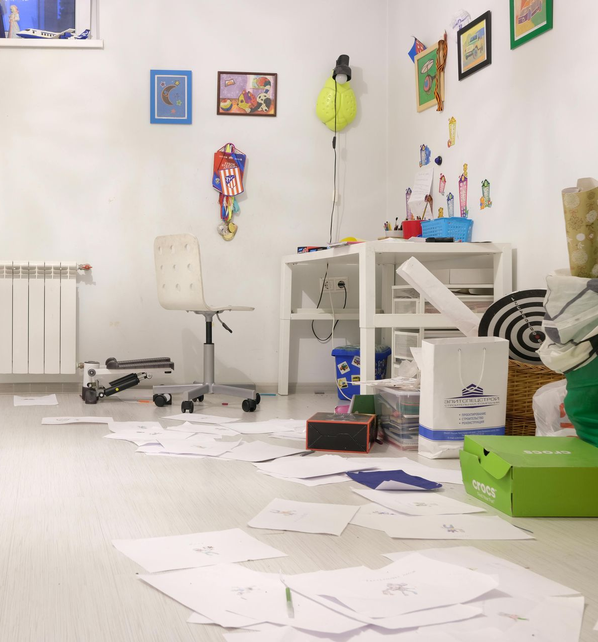 Messy room with scattered papers, a desk, chair, wall drawings, and supplies to clean up and trash out