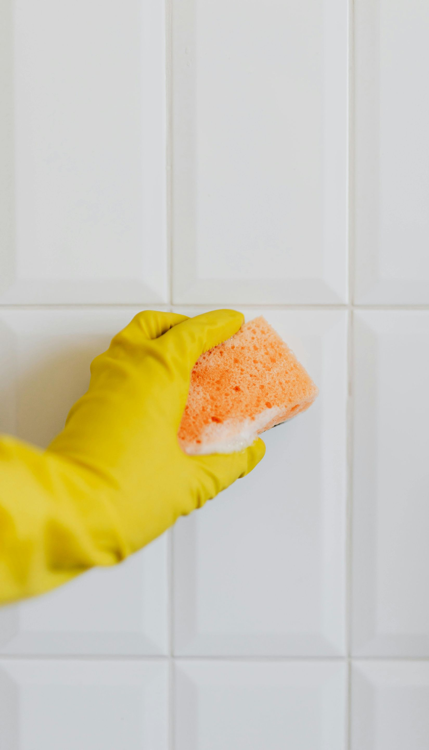 Closing Cleaners yellow glove with soapy orange sponge scrubbing white kitchen wall tiles