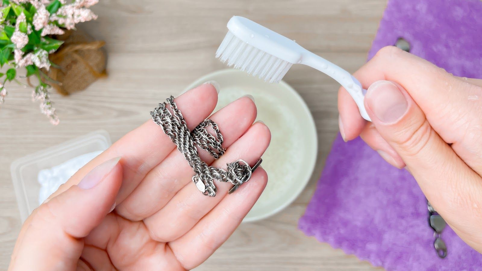 Hands cleaning a silver chain with a white brush over a tabletop with a bowl and purple cloth