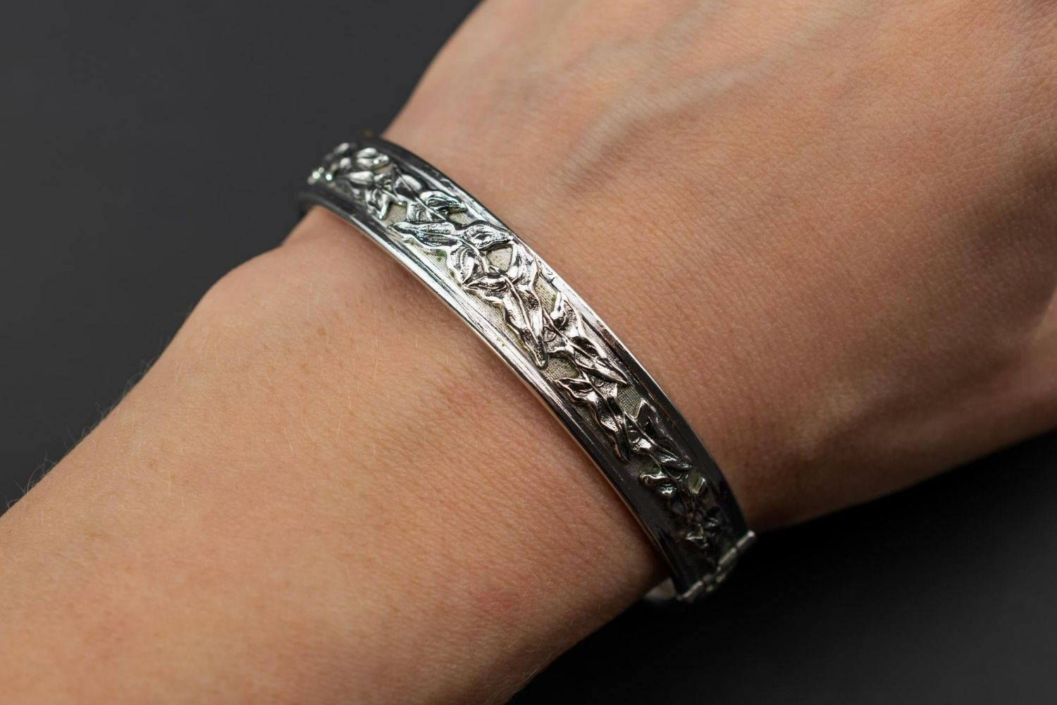 A silver bangle bracelet with an intricate embossed floral pattern worn on a wrist against a dark background.