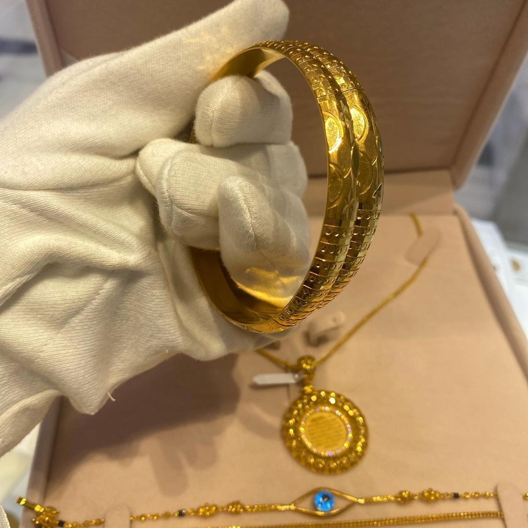 A person wearing a white glove is holding a gold bangle in front of a box of gold jewelry.