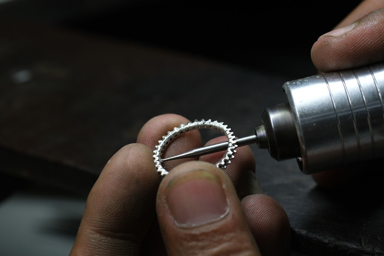 Hands holding a small gear ring near a rotary tool, working on precise metal jewelry or watch parts.