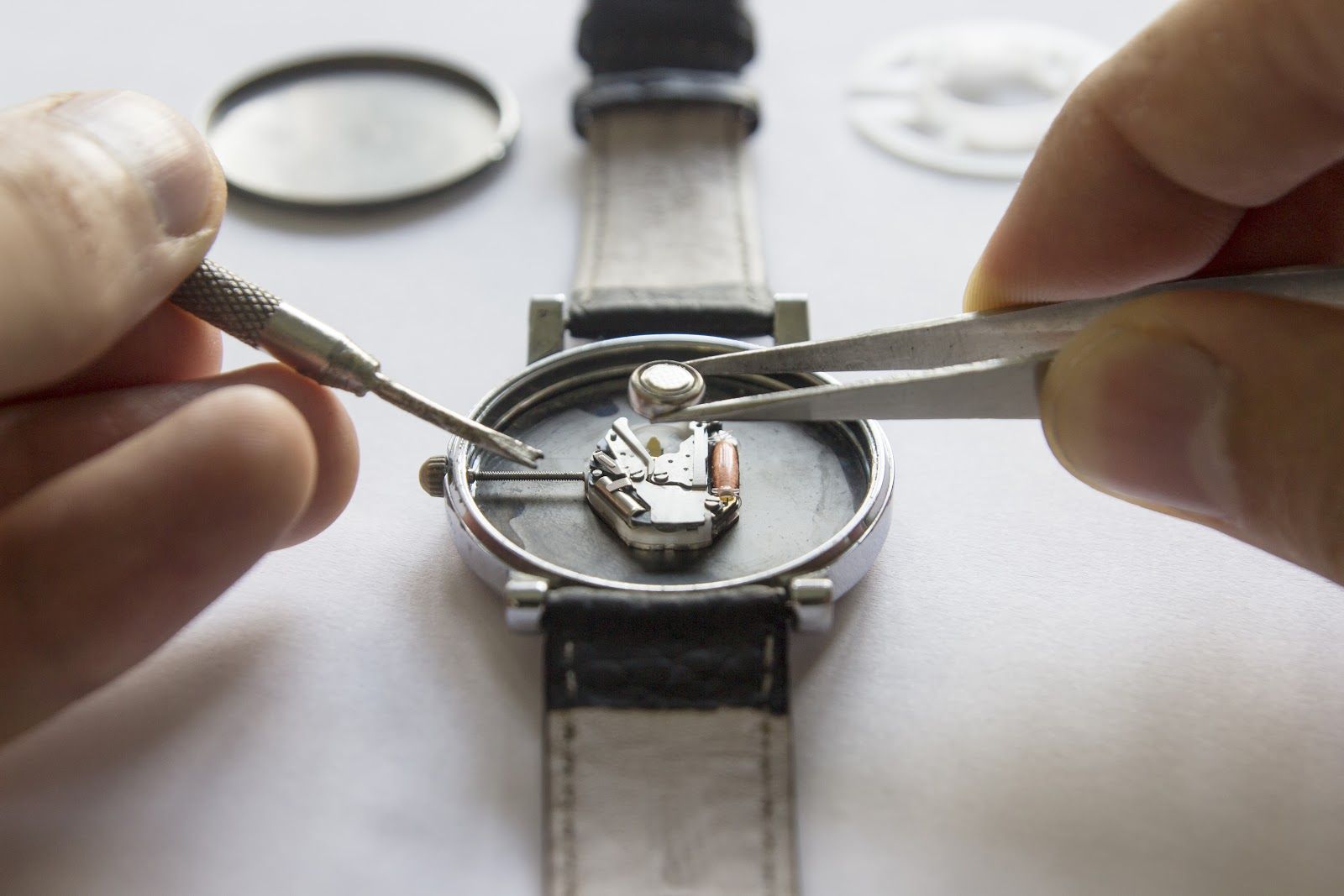 Hands using tweezers to repair a wristwatch movement on a white surface