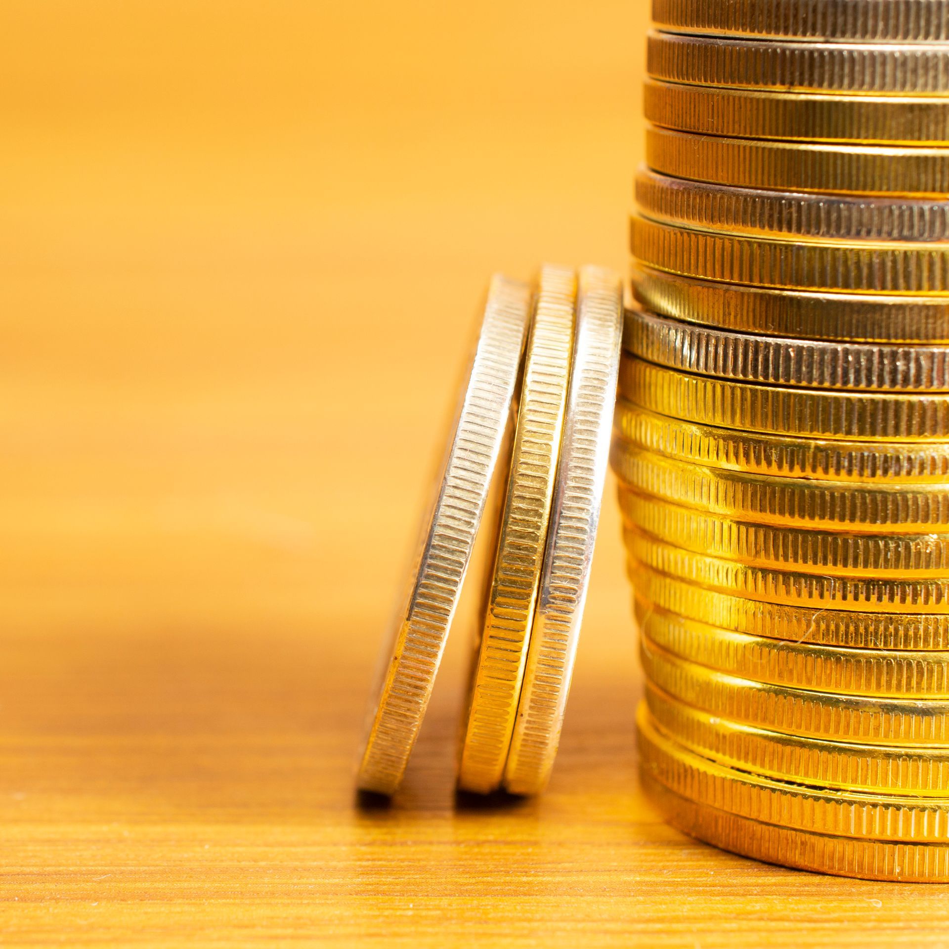 Two silver coins leaning beside a tall stack of gold coins on a warm yellow background
