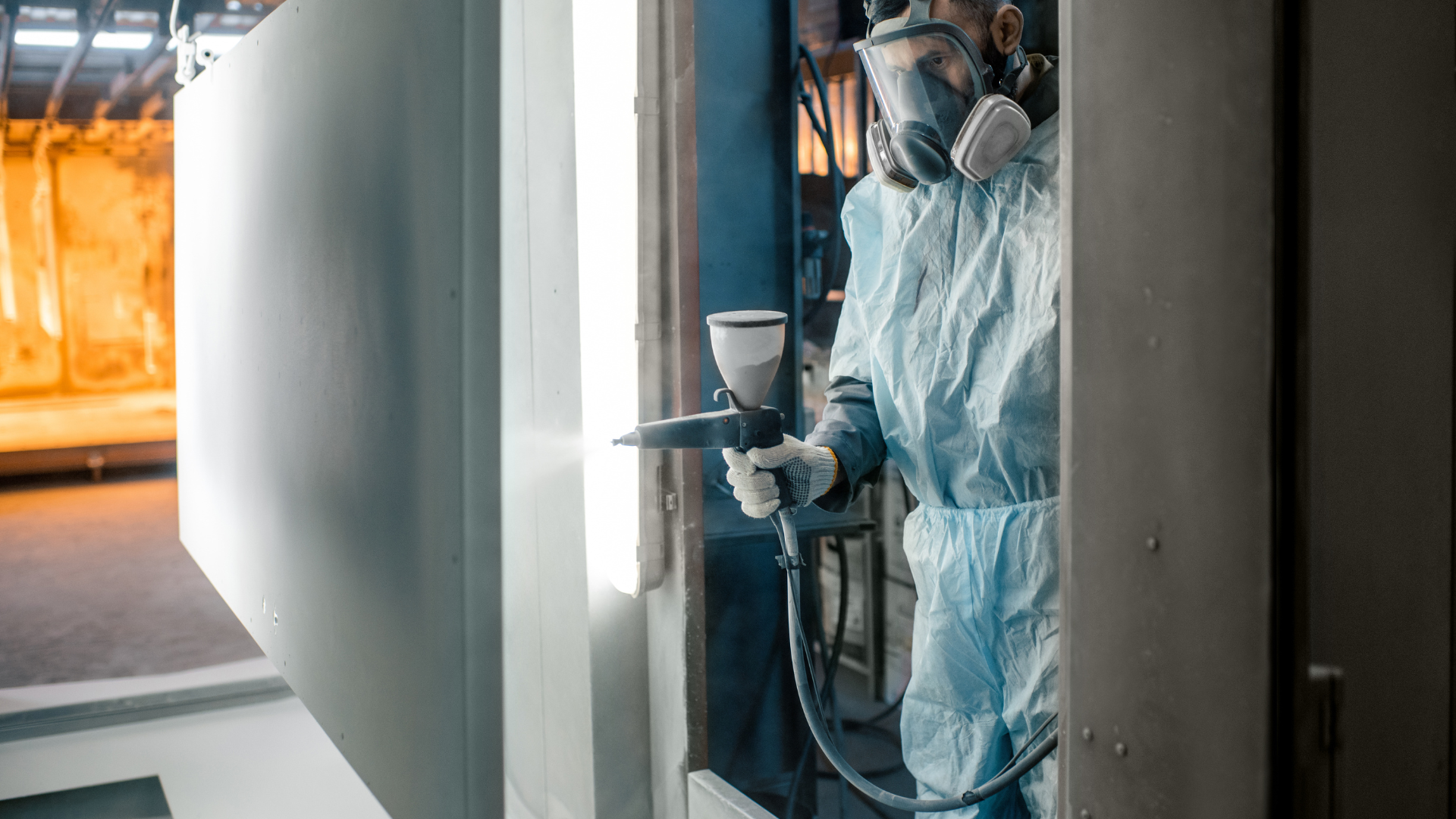 Worker in protective gear spray-painting a metal wall inside an industrial booth