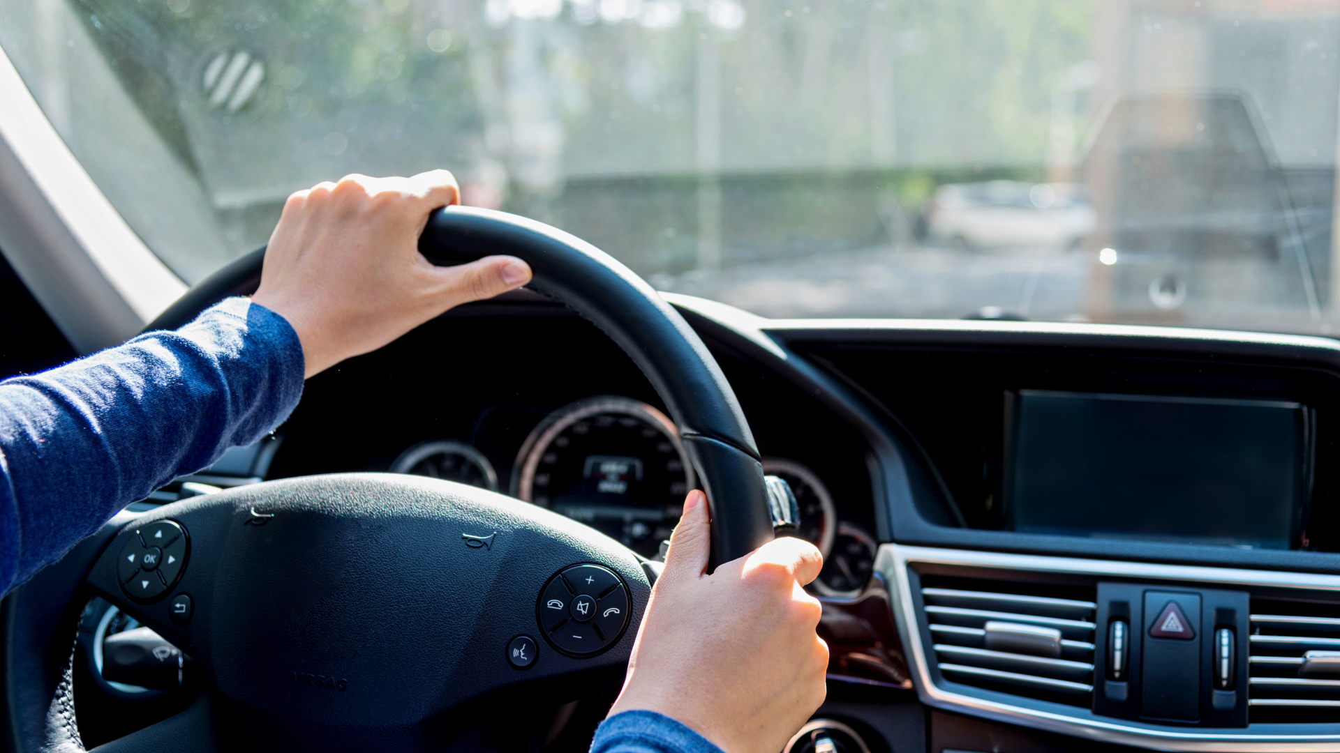 Driver’s hands on a steering wheel inside a car, dashboard and center console visible.