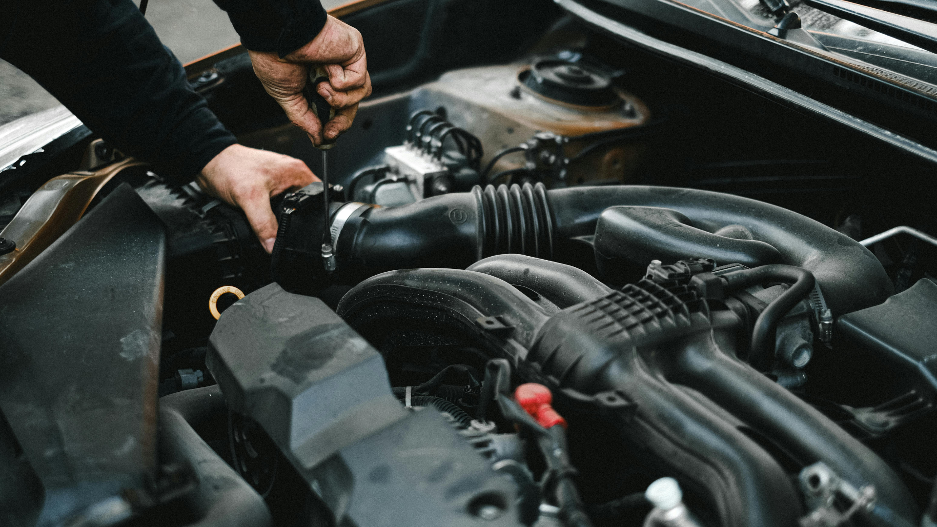 Hands working on a car engine in a garage, with hoses, tools, and engine components visible.