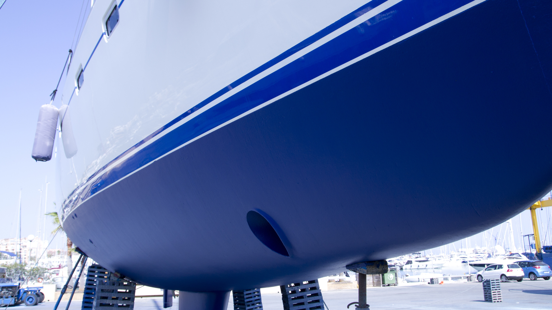 Close-up of a blue-and-white boat hull on a marina lift with railings and moored boats in the background
