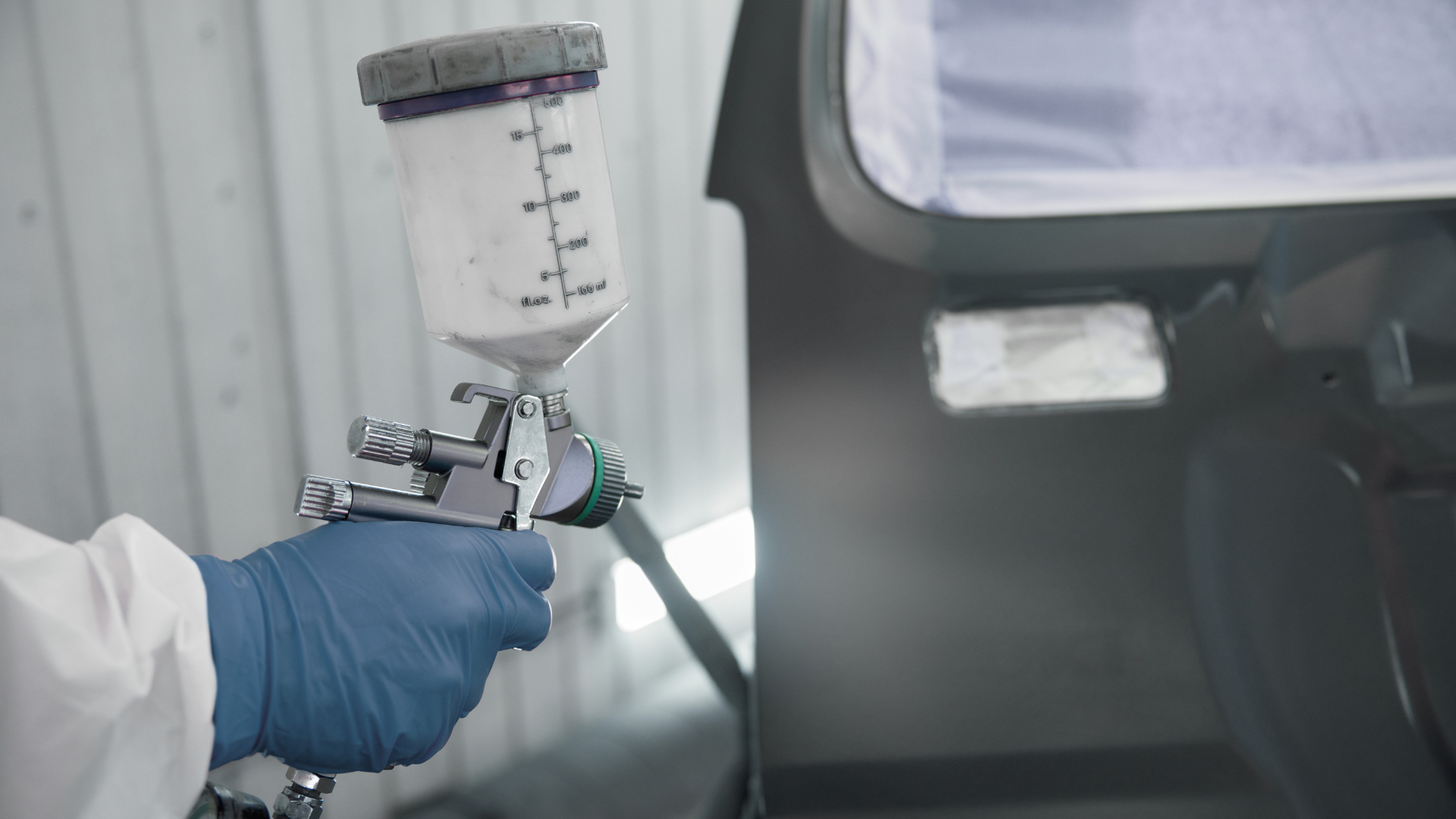 Gloved hand holding a spray gun with paint cup, spraying a car body in a workshop