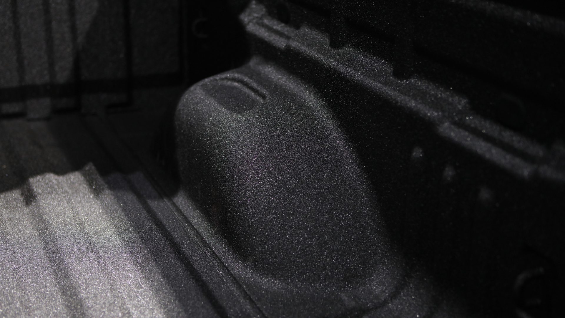 Black textured car seat or armrest in a dim interior, with a dark floor mat beside it.