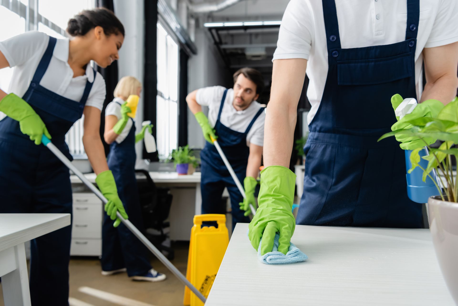 Quattro addetti alle pulizie professionisti in uniforme blu scuro e guanti verdi riordinano un ufficio, lavando i pavimenti e pulendo la superficie di una scrivania.