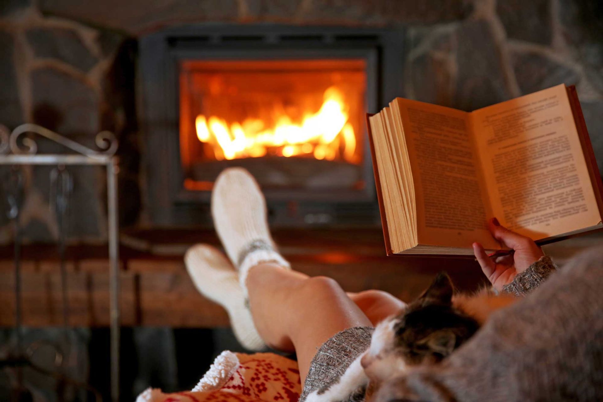 Person reading a book by a fireplace, cat on lap, feet in socks. Fire burning. Warm interior. Person reading a book by a cozy fireplace with a cat resting nearby