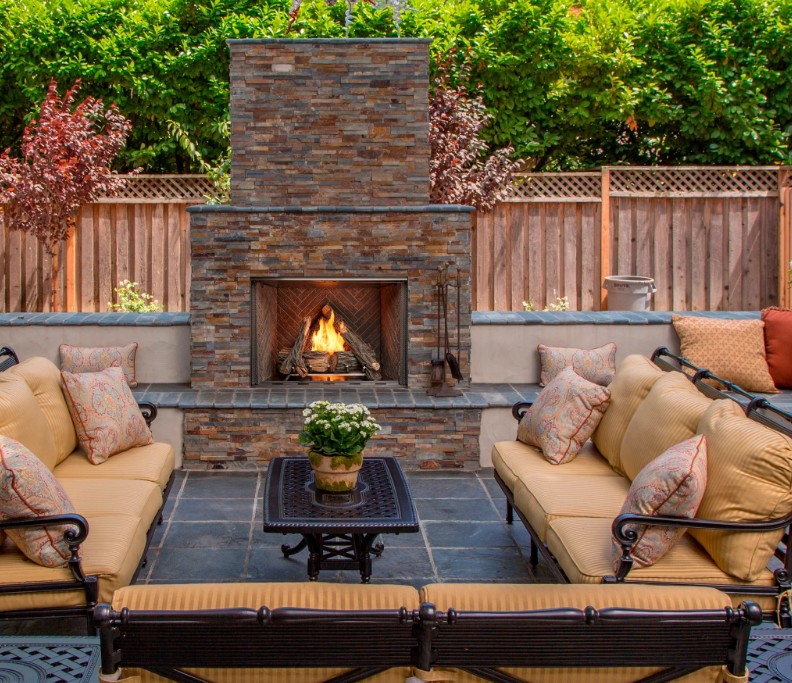 an outdoor patio seating area featuring a central stone fireplace with a burning fire.