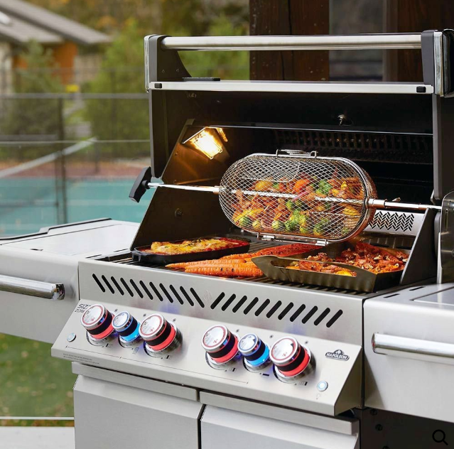 Outdoor kitchen with brick oven, grill, and countertop. Blue umbrella and fall foliage in the background. A stainless steel outdoor grill with a rotisserie basket filled with vegetables cooking above other food on the grates.