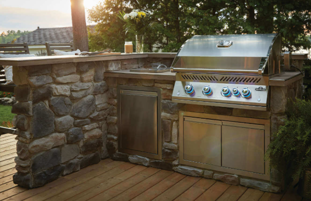 Outdoor kitchen with a built-in grill, stone counter, brick patio, and two bar stools. Outdoor kitchen with a stone-clad island, stainless steel grill, and cabinetry on a wooden deck at sunset.