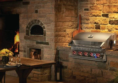 Man grilling food on outdoor kitchen, dining table with umbrella on patio overlooking lake. An outdoor kitchen with a stone pizza oven, a lit fireplace, a stainless steel grill, and a rustic wooden dining table.