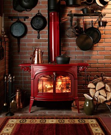 Vermont Castings Radiance Cozy brick hearth with a red wood stove, burning fire, hanging pans, and stacked firewood