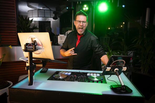 DJ in a black jacket and glasses, enthusiastically gesturing, behind a lit DJ setup outdoors.