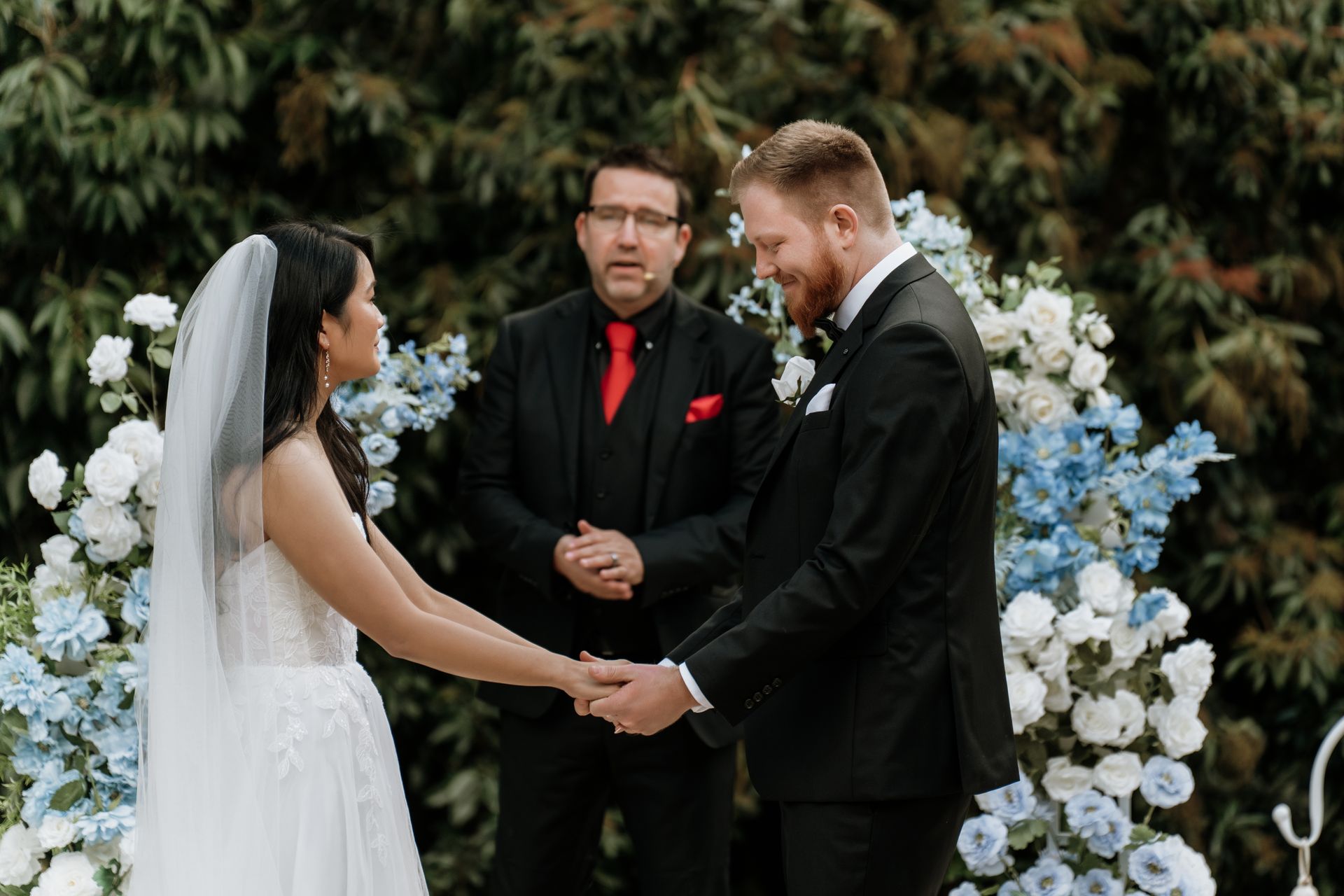 Bride and groom hold hands during an outdoor wedding ceremony, officiant stands beside them. Blue and white floral arch.
