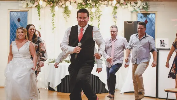 A bride and groom are dancing at a wedding reception while a man sings into a microphone.