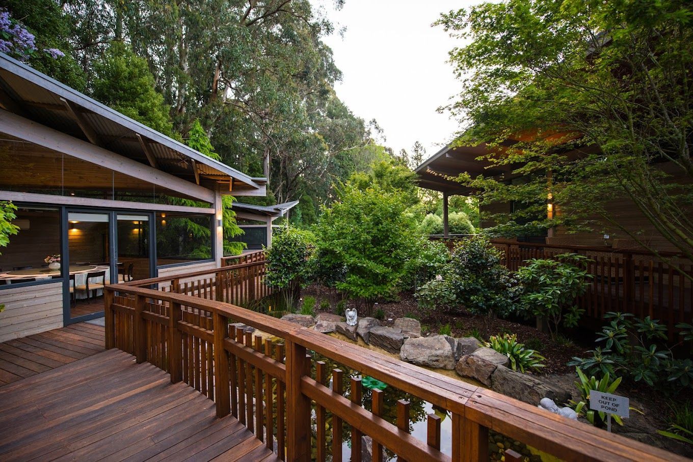 Wooden walkway with railings, leading to two buildings with dark roofs, surrounded by lush greenery and a pond.