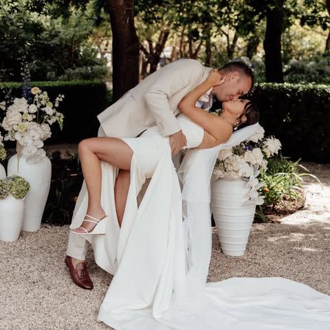 A couple in wedding attire kisses while dipping in an outdoor garden setting with large white floral vases.