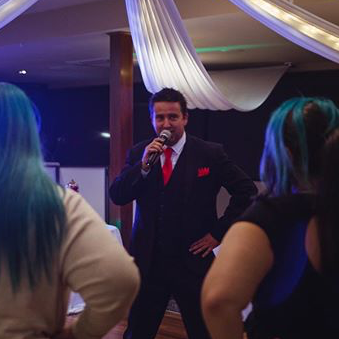 Man in suit, red tie sings into a microphone at an event with people.