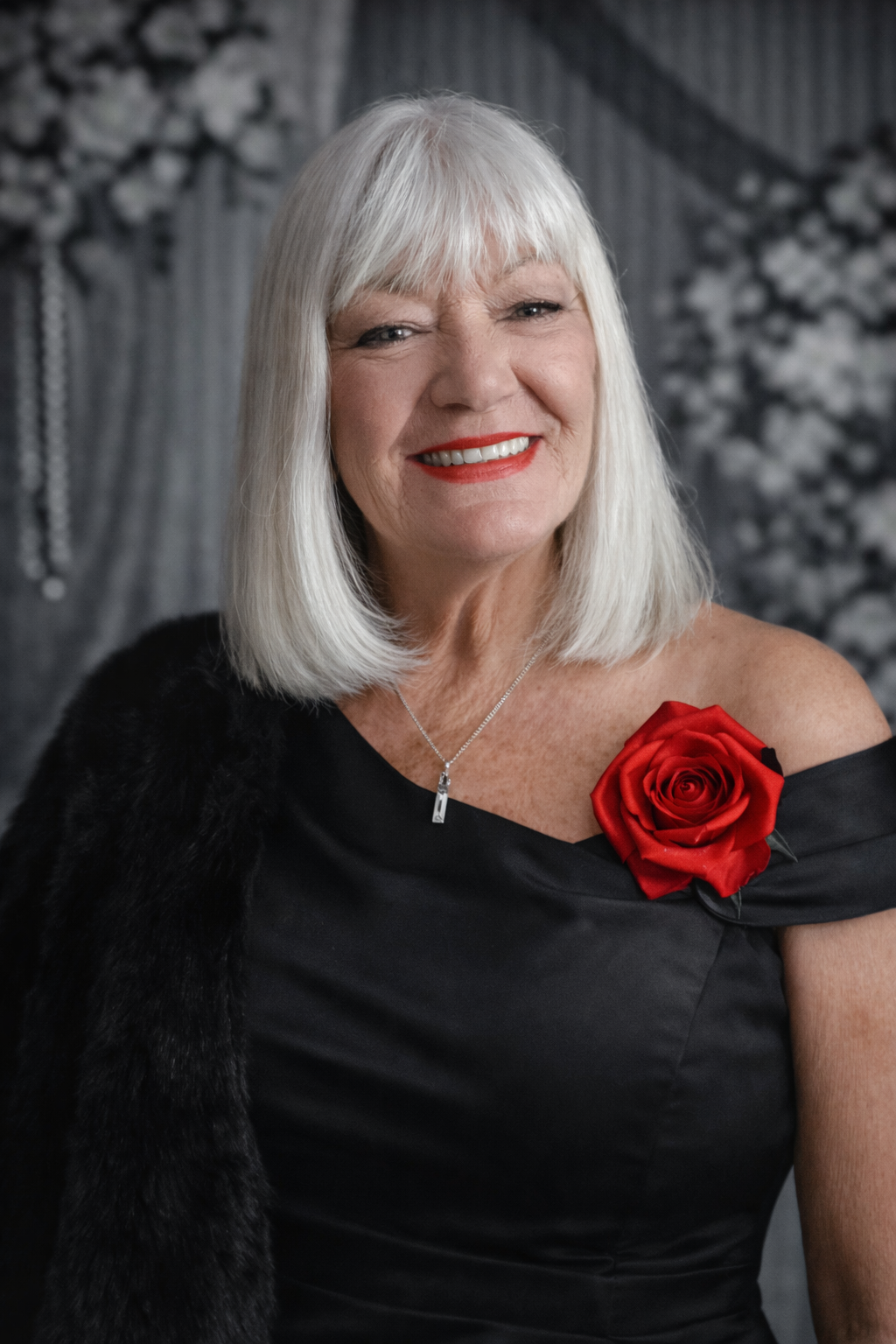 Woman with silver hair, wearing black dress with red rose, fur wrap, smiling.
