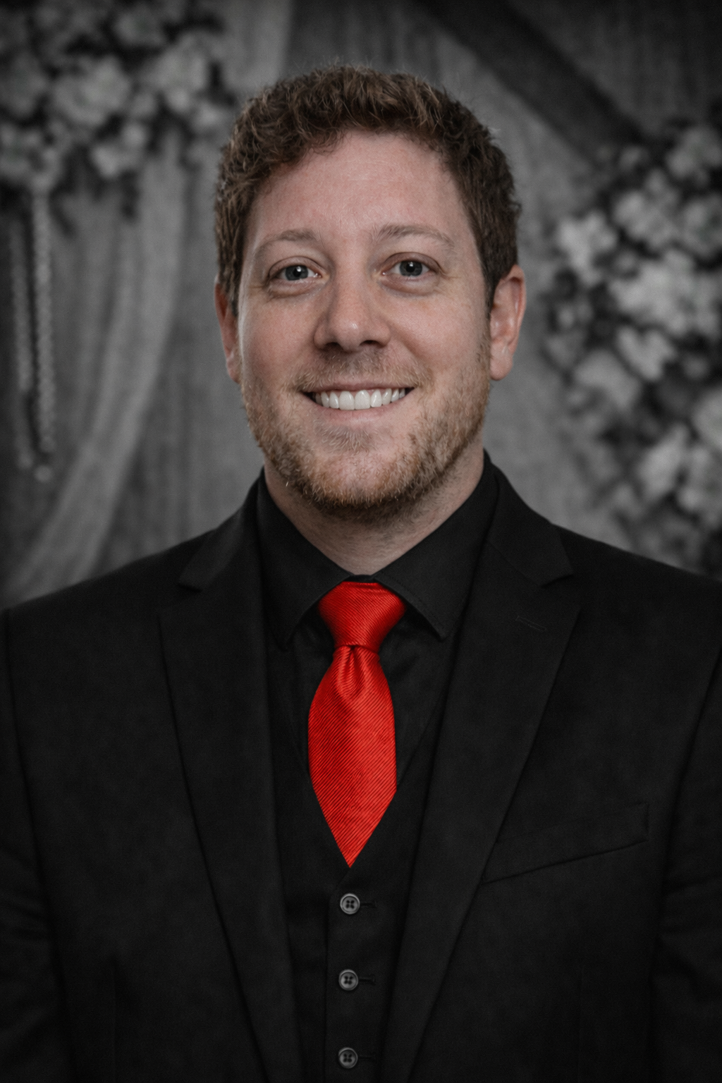 Man in a black suit with a red tie smiles at the camera against a faded background.