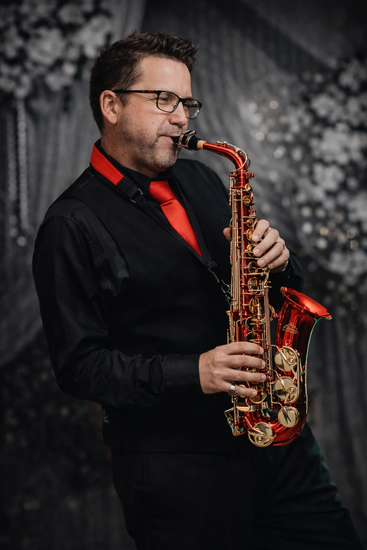 A musician playing a red alto saxophone, wearing a black shirt, vest, and a bright red necktie against a grey backdrop.