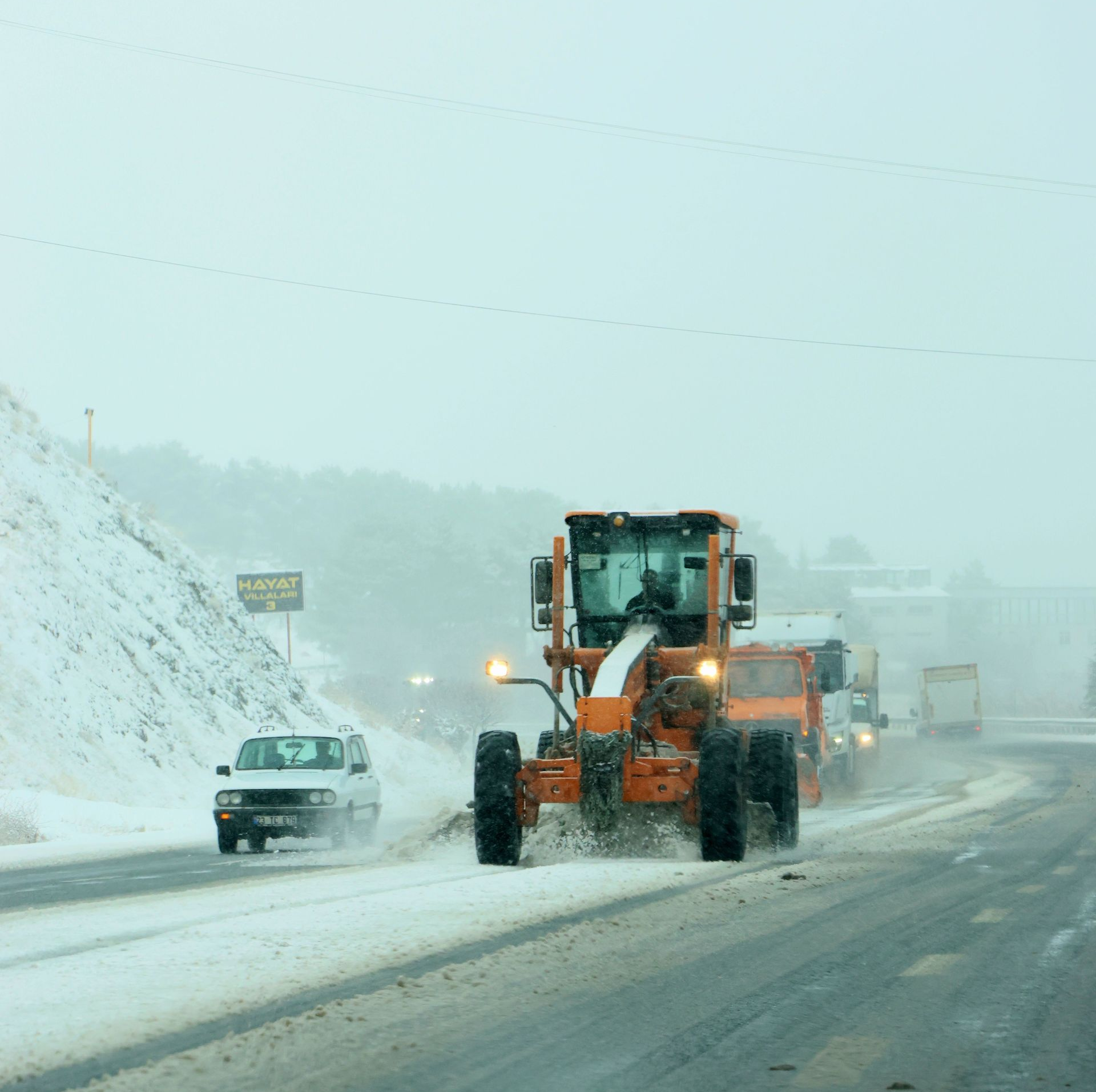 Orange snowplow clearing a snowy highway beside a parked car in heavy snowfall
