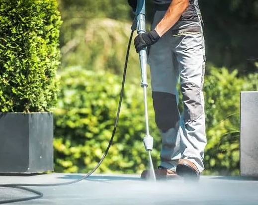 Worker pressure-washing pavement beside potted greenery outdoors