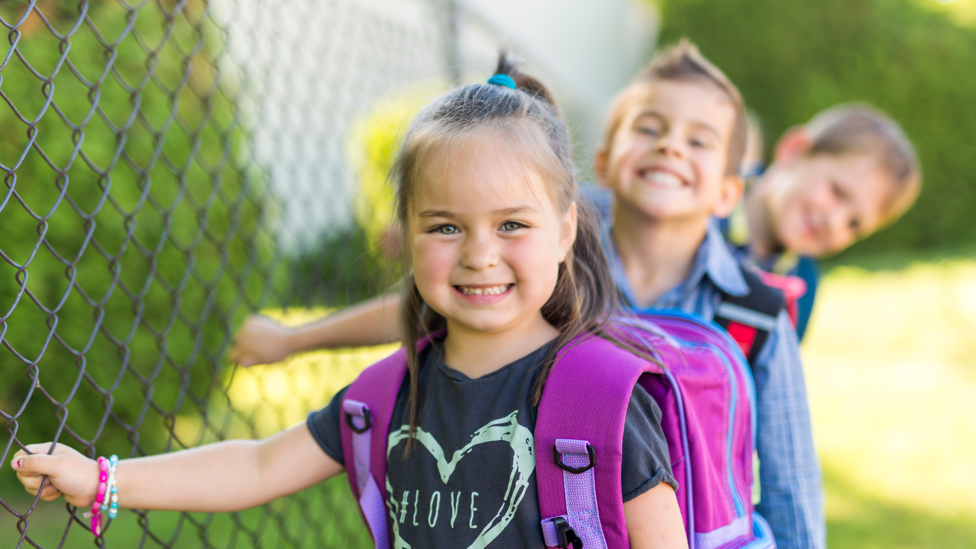 Three children with backpacks smile by a chain-link fence, outdoors.