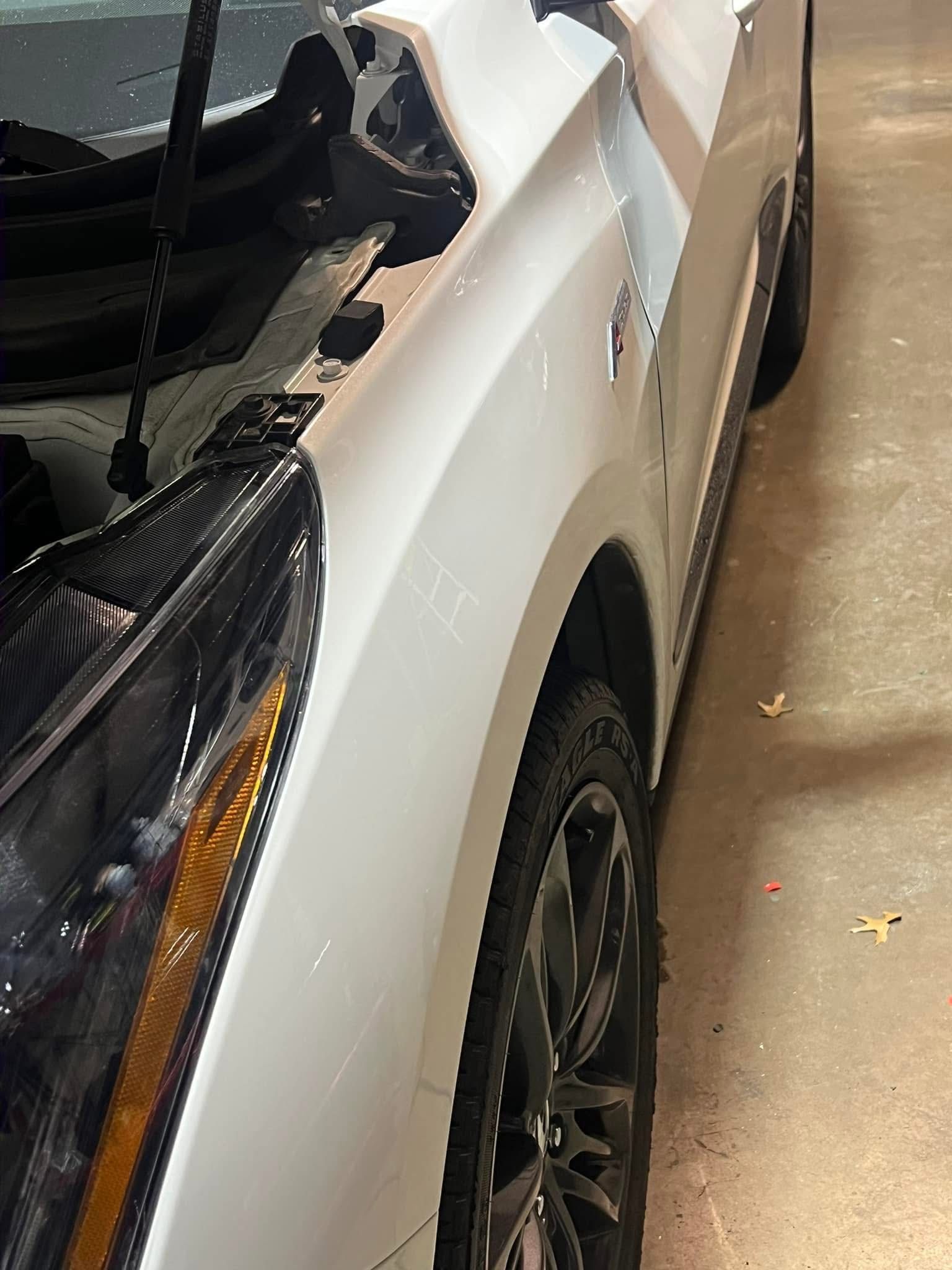 A close-up, high-angle shot of the front driver-side panel and wheel of a white car with the hood popped open.