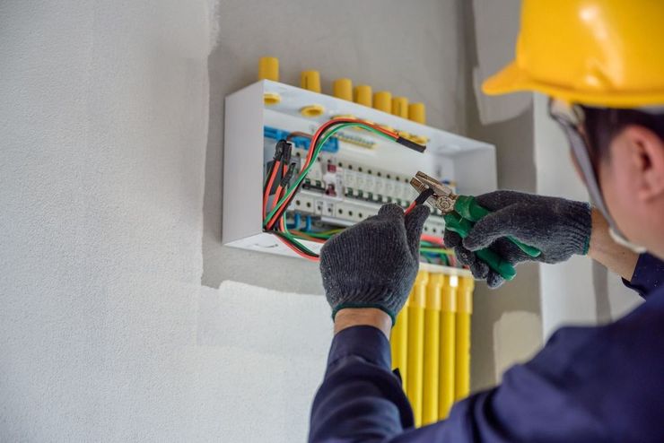 An electrician in a hard hat and gloves uses pliers to work on the wiring of an electrical panel on a white wall.